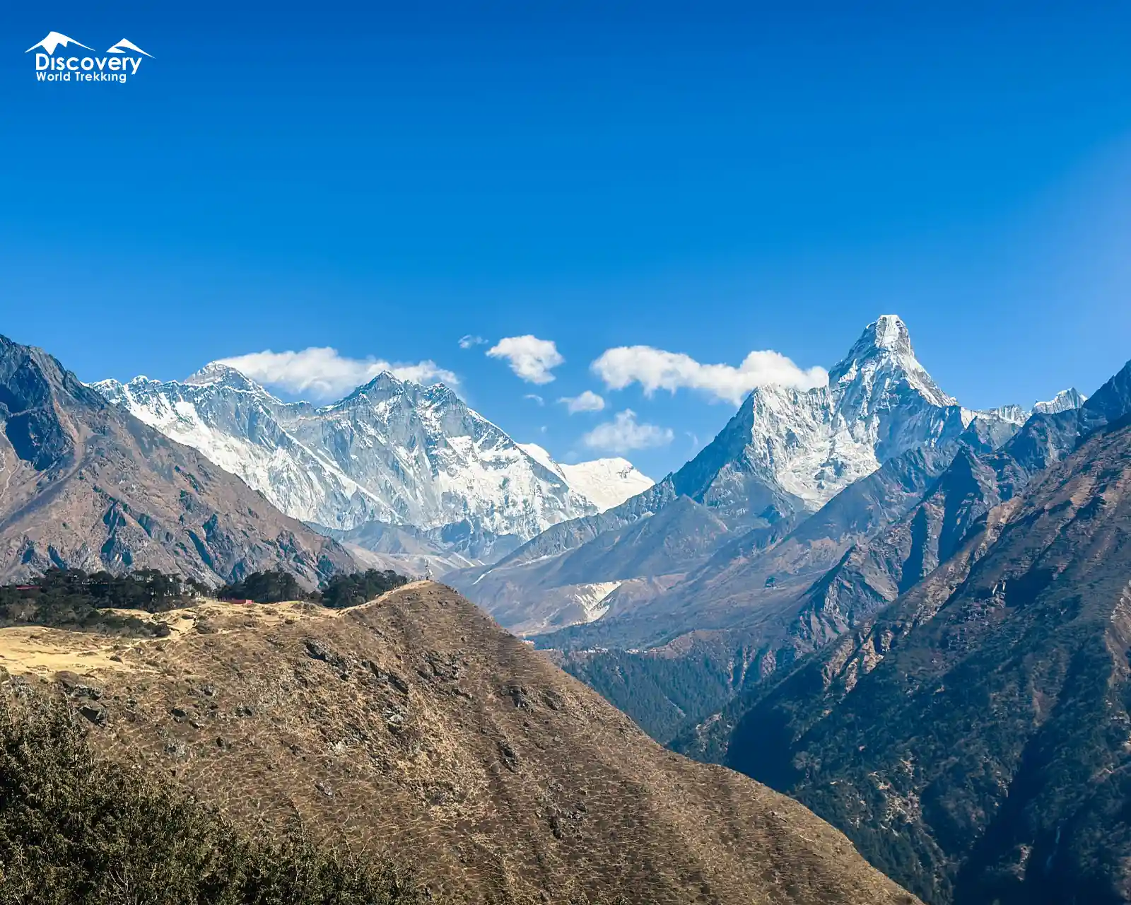 Panoramic view of Mount Everest (left) and iconic pyramid-shaped Ama Dablam (right) rising above rugged ridges under bright blue sky.