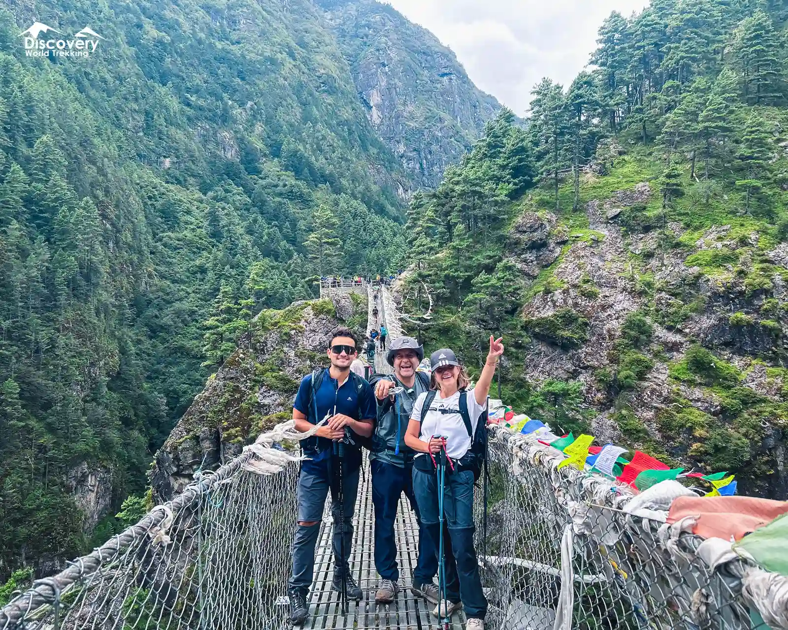 On Hillary Suspension Bridge over deep gorge, colorful prayer flags fluttering, lush green forests on steep hillsides in Everest region.